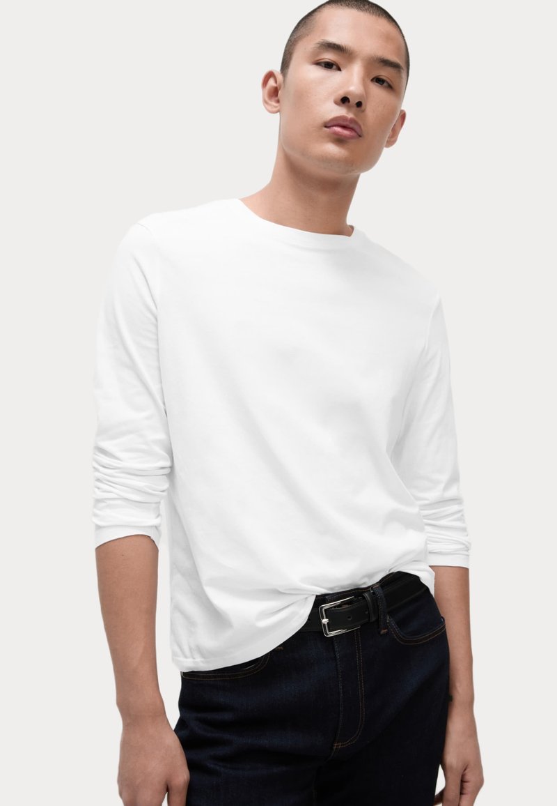 Langärmliges weißes T-Shirt aus Baumwolle mit Rundhalsausschnitt und enganliegenden Ärmeln, kombiniert mit dunklen Jeans und einem schwarzen Ledergürtel.