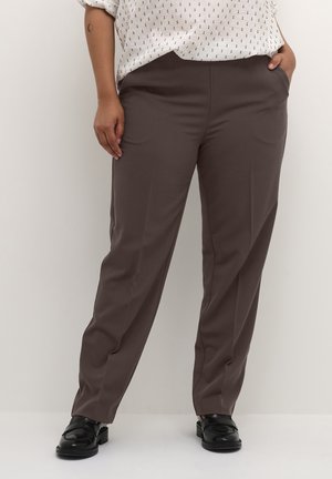Pantalon marron avec une texture lisse, doté de poches latérales et d'une coupe ajustée. Associé à un haut à motifs clairs et des chaussures noires.