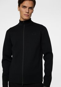 Svart zip-up tröja med hög krage, texturerat tyg och subtil logodetalj på bröstet. Modern, figursydd design med långa ärmar.