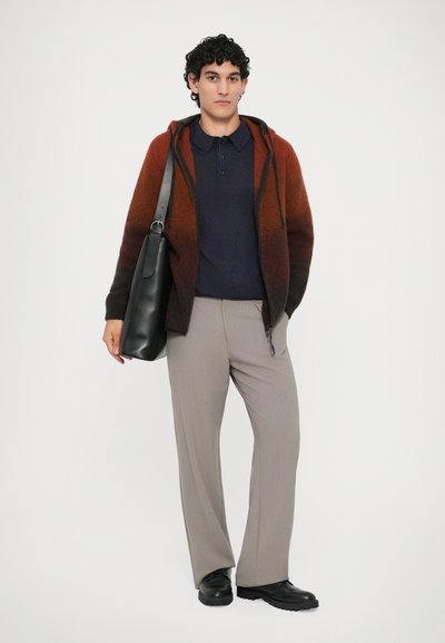 Jovem vestido com um hoodie em degradê de rust e preto, polo azul-marinho, calças bege, sapatos pretos e a carregar uma grande mala de couro preta.
