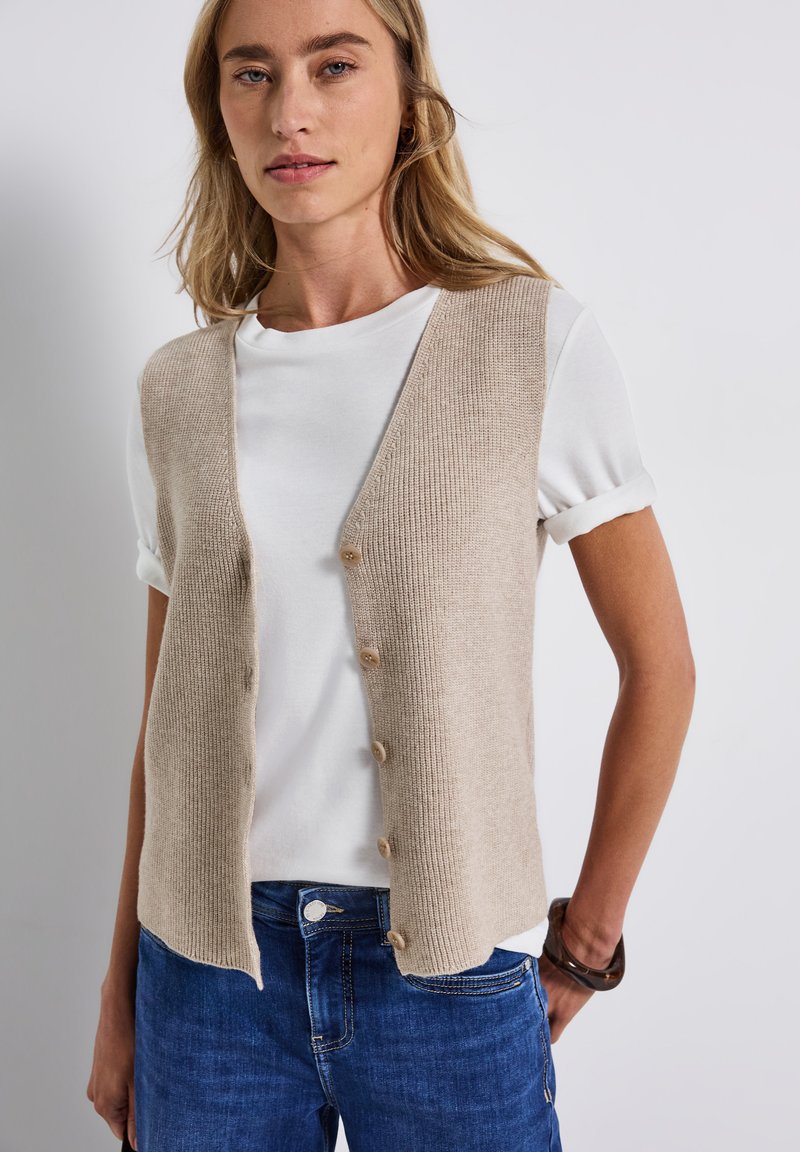 Chaleco de punto beige con escote en V y cinco botones, superpuesto a una camiseta blanca con mangas enrolladas, acompañado de unos jeans azules.