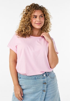 Femme souriante aux cheveux bouclés portant un t-shirt rose clair et une jupe en jean bleu clair boutonnée, posant devant un fond blanc uni.