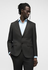 Άνδρας με σκουρόχρωμο δέρμα και ντραντλόκς, ντυμένος με σκούρο γκρι κοστούμι και ανοιχτό μπλε πουκάμισο, στέκεται με ένα χέρι στην τσέπη, κοιτάζοντας προς τα κάτω στο πλάι.