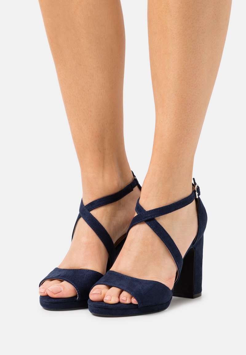 Pieds portant des sandales à talons hauts en daim bleu marine avec des brides croisées à la cheville et un design à bout ouvert sur fond blanc.