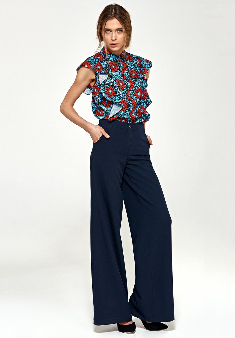 Bluzka w kwiatowy wzór w kolorze czerwonym, niebieskim i zielonym z falbankami na rękawach, zestawiona z spodniami o wysokim stanie i szerokich nogawkach w odcieniu granatowym, o gładkiej fakturze.