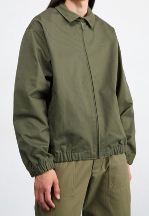 Buste masculin portant une veste zippée vert olive avec des poignets élastiques et un pantalon vert olive assorti avec de grandes poches avant.