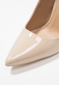 Beige lackläder högklackad sko med spetsig tå och en mjuk, glänsande yta. Inuti finns en mjuk beige foder.