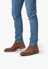 Des jambes portant un jean slim bleu et des bottes en cuir marron à lacets sur un fond blanc, avec un pied légèrement surélevé.