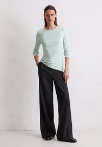 Hellgrünes Langarmshirt, kombiniert mit weiten, gestreiften schwarzen Hosen. Das Outfit zeichnet sich durch eine glatte Textur und eine lässige Passform aus.