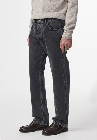 Jeans in denim nero con taglio dritto, caratterizzati da un design a cinque tasche e cuciture a contrasto, indossati con scarpe in pelle marrone.