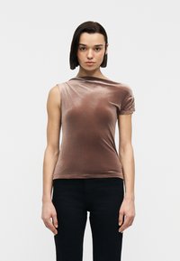 Jeune femme aux cheveux courts et foncés portant un haut en velours mauve asymétrique sans manches et un pantalon noir, debout devant un fond blanc uni.