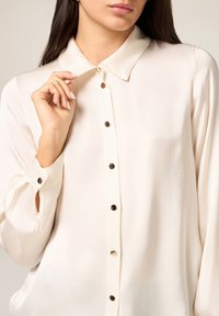 Crèmekleurige blouse met kraag, lange mouwen en knopen aan de voorkant. Voorzien van zwarte knopen en een zijdezachte textuur.