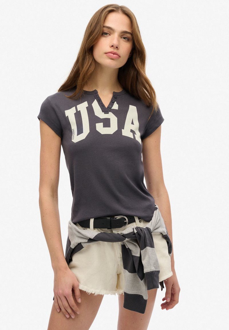 Mørk grå t-shirt med "USA" trykt i creme, korte ærmer, knaplukning ved halsen. Sammen med lyse shorts og en bundet stribet sweatshirt.