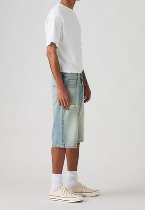 Shorts en denim bleu clair arrivant aux genoux avec des détails abîmés, associés à un t-shirt blanc à manches courtes et des baskets beiges.