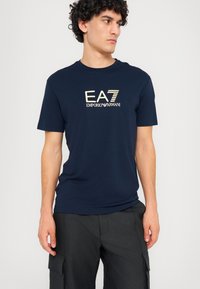 Tamsiai mėlynas medvilninis T-shirt su auksiniu logotipu "EA7 Emporio Armani" ant krūtinės. Trumpomis rankovėmis ir apvalia iškirpte. Lygi tekstūra.