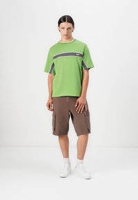 Green short-sleeve t-shirt with grey stripe and logo, paired with brown cargo shorts featuring pockets. White sneakers and visible socks.