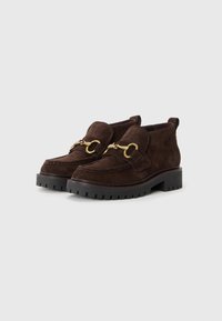 Mocassins en suède marron avec un bout rond, une chaîne en or décorative, et une semelle en caoutchouc noire avec un motif de tread texturé.