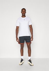 Hvid kortærmet t-shirt med sort logo, parret med sorte shorts med orange striber og sorte sneakers. Simpelt design, atletisk pasform.