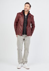 Rote Lederjacke mit hohem Kragen, über einer schwarzen Pufferweste getragen. Helle graue Jeans und weiße Sneakers runden den Look ab.