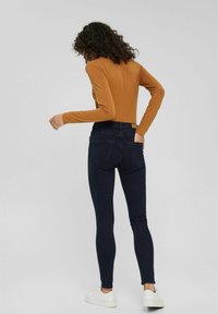 Kvinna med lockigt hår som bär en senapsgul långärmad tröja, mörka tajta jeans och vita sneakers, stående med en hand i bakfickan.