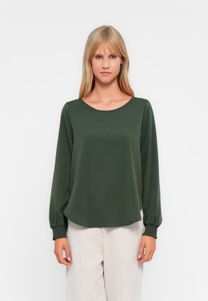 Blouse verte foncée à manches longues avec un col arrondi et des poignets froncés. Texture lisse, design fluide, associée à un pantalon de couleur claire.
