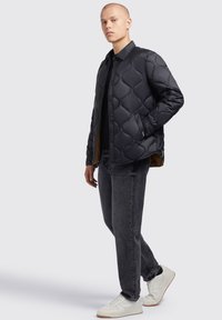 Sort quiltet jakke med knaplukning, der har en glat tekstur og rundet mønster; båret over en sort top og mørkegrå jeans.
