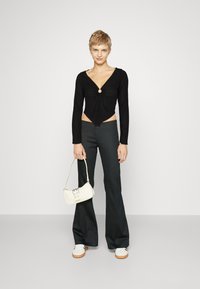 Svart langermet cropped topp med vridd frontknute og rund detalj, kombinert med høy midje svarte flared bukser og en liten hvit veske.