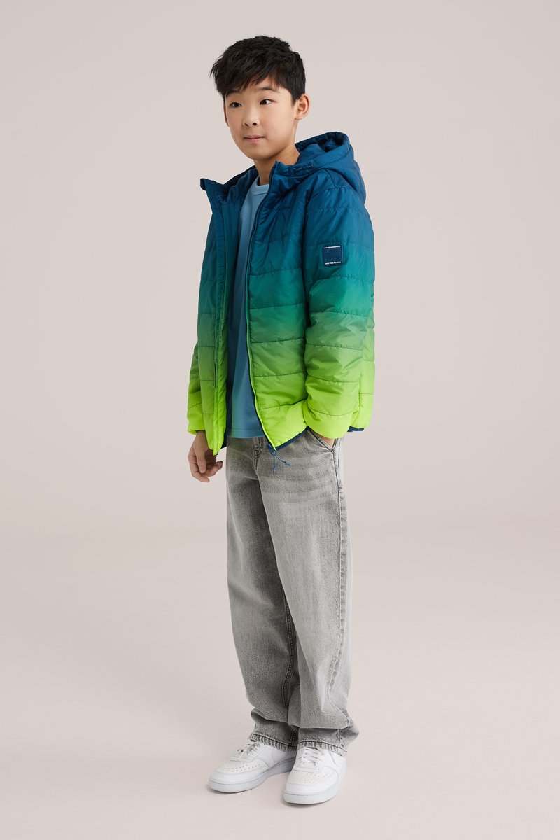 Chlapec stojaci v prechode bundy z modrej do zelenej, svetlo modrej košeli, sivých džínsoch a bielych teniskách na jednoduchom pozadí.
