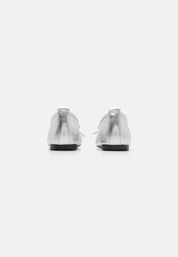 Ballerines métalliques argentées avec un bout arrondi, une semelle plate et un petit talon. Présentent une texture lisse et un détail lacé à l'avant.