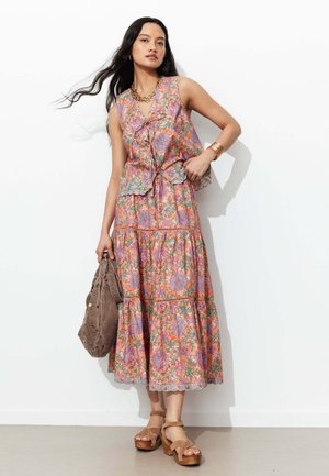 Femme en top sans manches fleuri et jupe midi assortie, tenant un sac en daim marron, portant des sandales à plateforme beige et des bijoux en or.