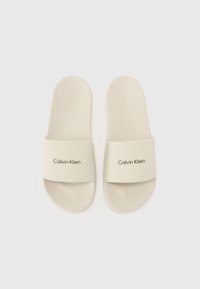 Beige glidsandaler med en bred stropp som har "Calvin Klein"-logoen. Teksturert fotseng og glatt overmateriale. Flat design.