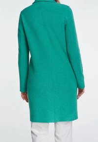 Femme portant un long manteau en laine teal vu de dos, associé à un pantalon blanc sur un fond uni.
