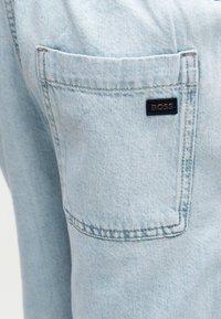 Τσέπη από ανοιχτό μπλε τζιν με ορατές ραφές και μια μικρή μαύρη ορθογώνια ετικέτα με την ένδειξη "BOSS" στο πάνω δεξιά μέρος.