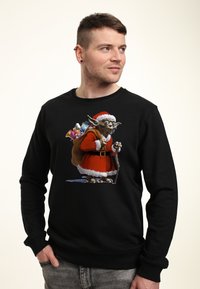 Svart sweatshirt med en detaljerad grafik av en varelse i en röd tomtedräkt med vit kant, som bär en väska med färgglada presenter.