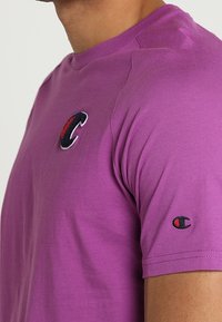 Fialové tričko s krátkým rukávem vyrobené z bavlny, s vyšitým logem na prsou a menším logem na rukávu.