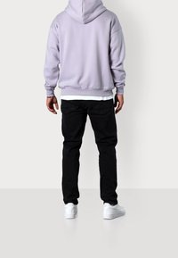 Felpa con cappuccio di un delicato colore viola chiaro, dal tessuto morbido, abbinata a jeans neri e sneaker bianche, vista da dietro.