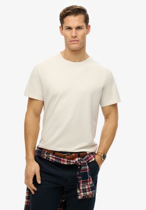 Camiseta de algodón beige con cuello redondo, mangas cortas, combinada con pantalones azul marino y una camisa de cuadros atada a la cintura, que tiene un cinturón marrón.