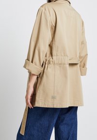 Beige casual jack met opgerolde mouwen, een trekkoord in de taille en een klein logo tag aan de achterkant, gecombineerd met losse blauwe broek.