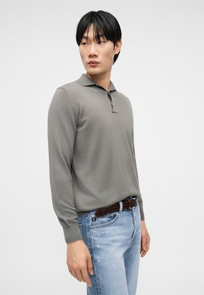 Νέος άνδρας που φορά γκρι μπλουζάκι πόλο με μακριά μανίκια και ανοιχτόχρωμα μπλε τζιν με καφέ πλεκτή ζώνη, κοιτάζοντας πλάγια.