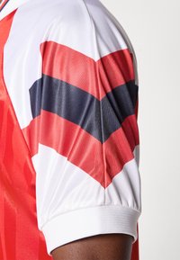 Camisola vermelha com detalhes em listras brancas e pretas nas mangas. Feita de um tecido suave com punhos brancos canelados.