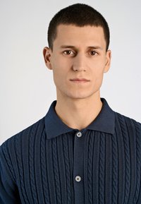 Tumedat sinise värvusega kudumipolo särk, millel on tekstuurne keraamika muster, kraega disain ning kaks nööpi kaela juures.