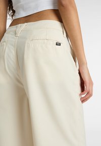 Pantaloni a gamba larga di colore crema, realizzati in tessuto morbido, con tasche posteriori e un piccolo logo black Vans.