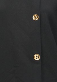 Μαύρο υφασμάτινο πουκάμισο με χρυσά κυκλικά κουμπιά που φέρουν ανάγλυφα γράμματα "A" και "R" στο μπροστινό μέρος, με λεία υφή.