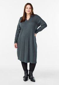 Kleita ar garām piedurknēm, V-veida kakla izgriezumu tumši zaļā krāsā ar teksturētu stieļu adījumu. Saskaņota ar melnām potīšu zābakiem un zeķubiksēm.