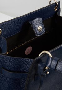 Navy nahka handbag, jossa on teksturoitu pinta, kultasävyiset metalliosat, sisäinen vetoketjullinen tasku ja merkkilaatta. Avautuu laajasti helppoa pääsyä varten.