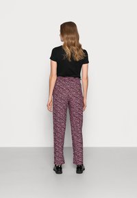 Woman with long wavy hair wearing a black short-sleeve top, high-waisted purple patterned pants, and black shoes, standing facing away.