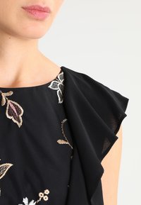 Czarna bluzka z kwiatowym haftem, z okrągłym dekoltem i falbaniaste krótkie rękawy. Miękka faktura, skomplikowane wzory kwiatowe w kolorze kremowym i różowym.
