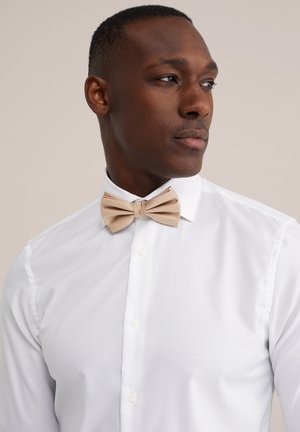 Uomo con camicia bianca e papillon beige, che guarda di lato su uno sfondo neutro e uniforme.