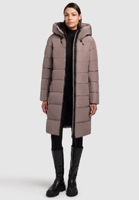 Długa pikowana kurtka w jasnym taupe z kapturem, zapinana na zamek błyskawiczny z przodu, z elastycznymi wstawkami i dwoma bocznymi kieszeniami. Noszona na czarnym ubraniu.