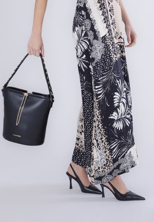 Mujer con una falda larga estampada, tacones slingback negros, sosteniendo un bolso tipo cubo negro con detalle de cadena dorada.